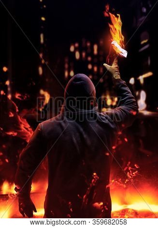 Photo of a protester in mask and hood holding and raising up burning molotov cocktail on night streets city background.