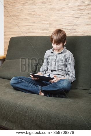 Lonely boy sittng on sofa with tablet computer stuck at home during isolation. Quarantine and lockdown are protective measures against spreading of coronavirus pandemic disease