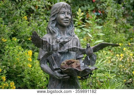 Statue Of A Girl With Birds On A Background Of Trees And Flowers