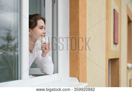 Caucasian Young Woman Smiles, Looks Out Of The Apartment Window And Takes Off Her Protective Mask. E