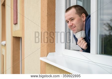 Caucasian Adult Man Smiles, Looks Out Of Apartment Window And Takes Off Protective Mask. End Of Quar