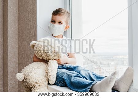 A Small Child In A Mask Sits At Home In Quarantine And Looks Out The Window In A Place With A Teddy