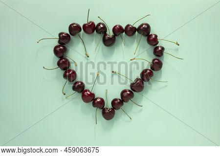 Red Cherries With Stems Lay Flat Closeup In A Heart Shape On Aqua Blue Background