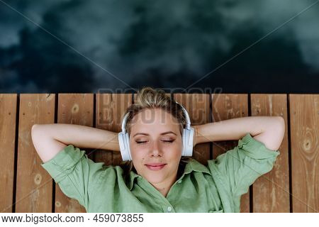 Rlaxed Woman Wearing Headphones Listening To Music Lying On A Pier By Natureal Lake In Summer