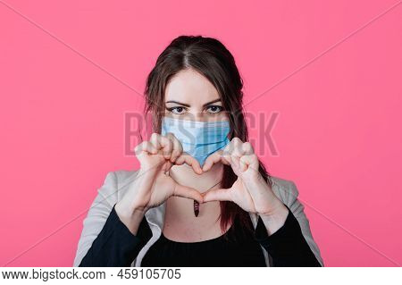 Young Beautiful Woman In Face Medical Mask Shows Heart Jesture, Pink Background