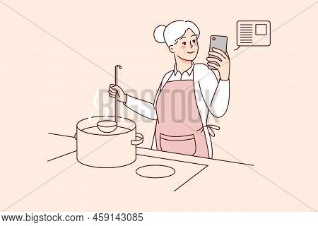 Elderly Woman Cooking Soup At Kitchen With Online Recipe On Cellphone. Smiling Modern Old Grandmothe