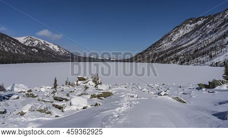 The Surface Of The Frozen Lake Is Covered With Clean Fresh Snow. Footprints Among The Boulders. Surr