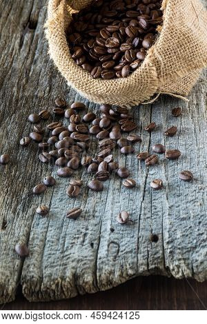 Coffee Beans In A Burlap Bag With Many Poured Out Onto A Rustic Wooden Table.