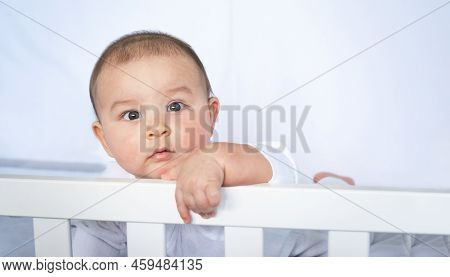 Cute Portrait Of A Baby In A Crib Close-up. A Child In White Clothes On White Underwear. Tenderness 