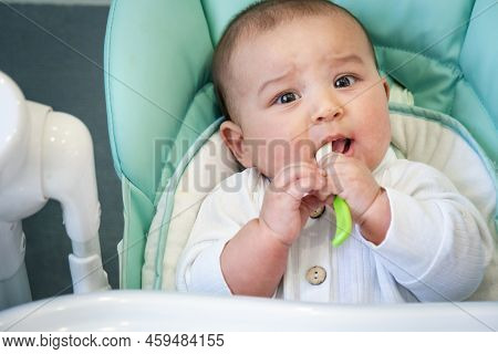 A Hungry Baby Is Gnawing On A Plastic Spoon At The Table On A High Chair. Teething, Whims, Itchy Gum