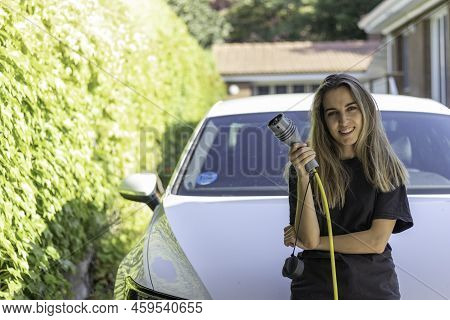 Portrait Of Youg Woman Looking And Smiling To Camera With An Electric White Car About To Be Pluged A