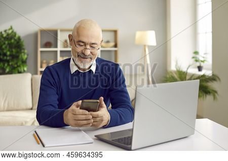 Happy Senior Man Using His Mobile Phone While Sitting At Desk With Laptop Computer