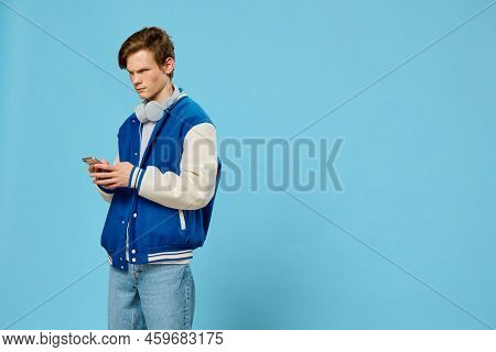 Young Stylish Guy In A Bomber And Jeans Stands With A Phone In His Hands And Headphones Around His N