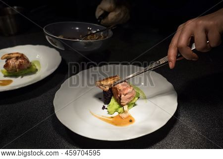 Chief Cook Putting Grilled Meat Piece Using Tweezers On Green Vegetables Puree White Plate With Sauc