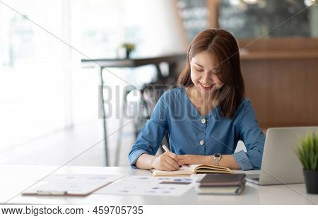 Smiling Pretty Girl Working On Laptop Computer While Sitting At Office Table, Taking Notes In A Diar