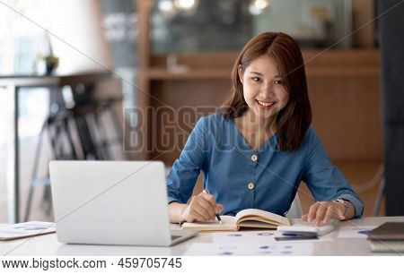 Smiling Pretty Girl Working On Laptop Computer While Sitting At Office Table, Taking Notes In A Diar