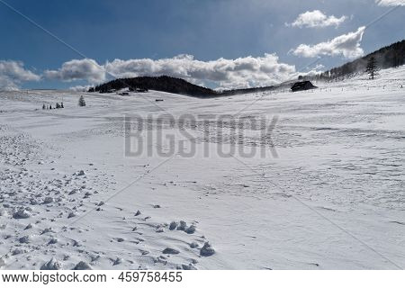 An Alpine Pasture Covered With Snow With Some Small Huts. The Sky Is Blue With A Few Clouds And The 