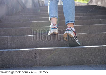 Close-up Of Woman Legs In Jeans And Sneakers Going Up Steep Stairs. Female Tourist In Casual Clothes