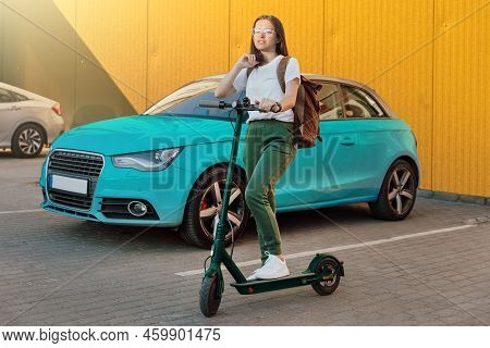 Young Woman In White Sneakers Standing On Electric Scooter In Front Of The Blue Car