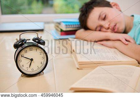 Focus On A Black Alarm Clock On Blurred Background Of A Tired Sleepy Schoolboy Falling Asleep On A T