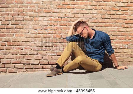 alkalmi fiatal ember ül a járda mellett egy téglafal a kezét a feje, és keres 