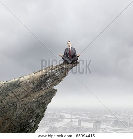 Young businessman sitting lotus pose on edge of rock mountain