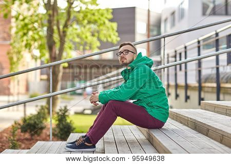 people, style, leisure and lifestyle - happy young hipster man in eyeglasses and jacket sitting on stairs in city