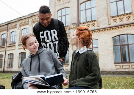 Multiethnic group of happy young people reading book and talking outdoors at the unversity campus