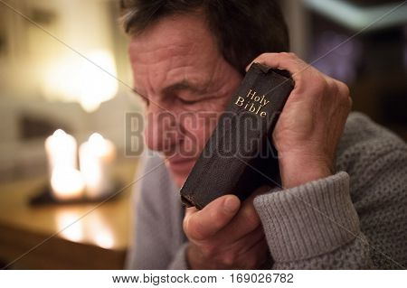 Senior man praying, holding an old Bible in his hands, eyes closed. Burning candles behind him. Close up.