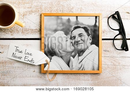 Black-and-white photo of senior couple in love in brown picture frame. Eyeglasses and cup of coffee laid on table. Studio shot on white wooden background.