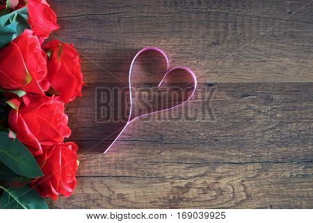 heart make from pink ribbin near a bloom red roses on wooden table for anniversary; valentine; congratulation; tell to love concept