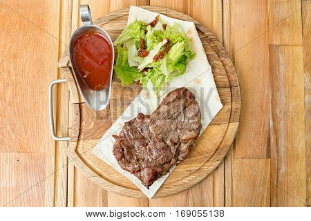cooked steak on a pita a wooden board standing next to a gravy boat with ketchup and greens
