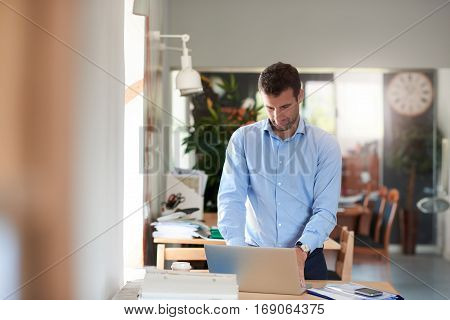 Confident and successful businessman wearing an open collar shirt standing at his desk and working on a laptop in his modern office