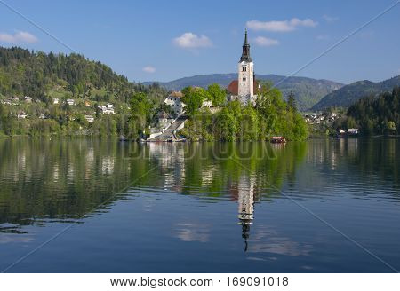 Island on Bled lake and pilgrimage church of the Assumption of Mary with mountain landscape background reflected in water at sunny day, Slovenia