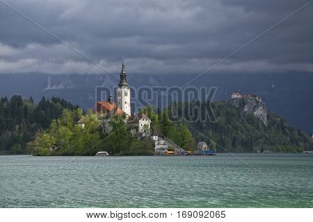 Island on Bled lake and pilgrimage church of the Assumption of Mary in sunlit with mountain covered dark clouds on background , Slovenia
