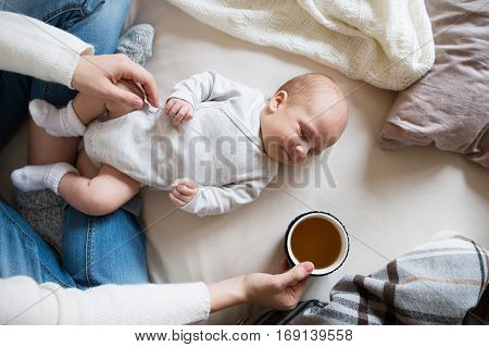 Unrecognizable mother holding a cup of tea and her newborn baby son lying on bed next to her