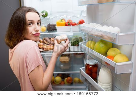 Young woman in the sleepwear eating sweet donuts near the refrigerator