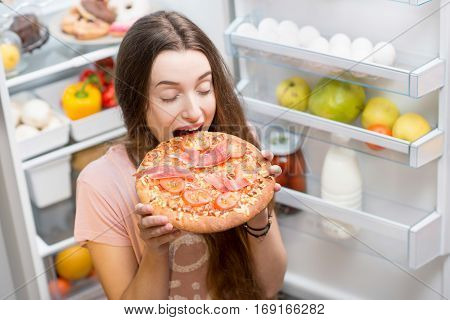 Young woman with pizza standing near the open refrigerator full of fruits and vegetales