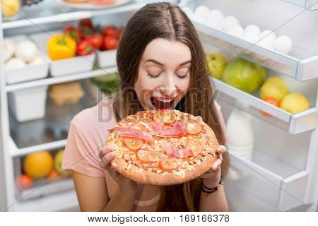 Young woman with pizza standing near the open refrigerator full of fruits and vegetales