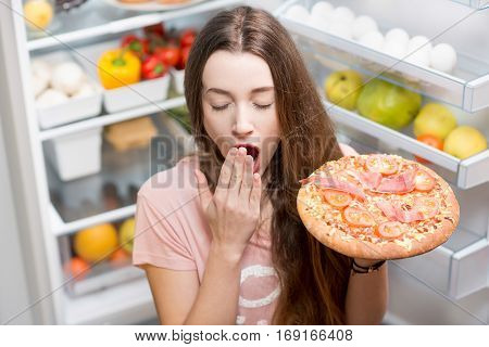 Young woman with pizza yawning near the open refrigerator full of fruits and vegetales