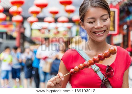 Chinese woman eating Bing Tang Hulu, a traditional chinese food snack from Beijing. Fruit candy stick sold in outdoor market place on Wangfujing street. Candied fruits on a bamboo stick.