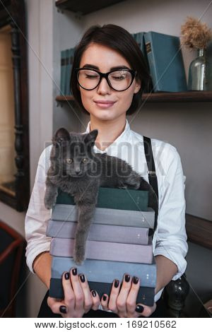 Vertical image of Smiling Authoress in glasses and white shirt with closed eyes holding books with cat on them