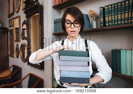 Authoress in glasses and white shirt holding books and standing near the bookshelf