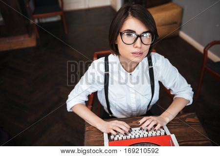 Top view of Authoress in glasses and white shirt sitting by the table, using typewriting and looking at camera