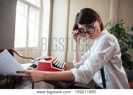 Side view of pretty authoress in glasses and white shirt sitting by the table with paper and typewriter and looking at camera