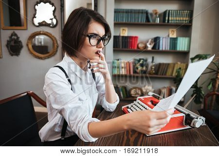 Side view of thoughtful authoress in glasses and white shirt sitting by the table with paper and typewriter