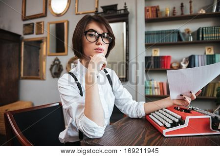 Side view of authoress in glasses and white shirt sitting by the table with paper and typewriter and looking at camera