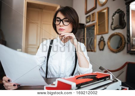 Authoress in glasses and white shirt sitting by the table with paper and typewriter