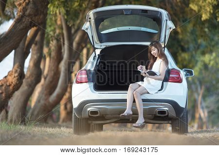 Woman relaxing outdoors looking happy and reading a book on back white car,green park view
