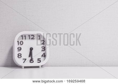 Closeup white clock for decorate show half past six or 6:30 a.m. on white wood desk and cream wallpaper textured background in black and white tone with copy space